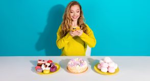 Junge Frau vor türkisem Hintergrund fotografiert mit ihrem Smartphone drei Teller mit Süßigkeiten, Kuchen und Gebäck, die vor ihr auf einem Tisch stehen.