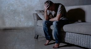 Ein Mann sitzt in einem abgedunkelten Raum auf einem Sofa, stützt den Kopf in die Hand und blickt nach unten.