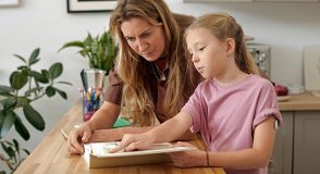 Mutter und Tochter sitzen gemeinsam am Tisch und betrachten ein Buch. Am Oberarm des Mädchens ist ein Glukosesensor zu sehen; die Szene steht für die gemeinsame Bewältigung des Diabetes-Alltags bei etwaigen weiteren Einschränkungen.