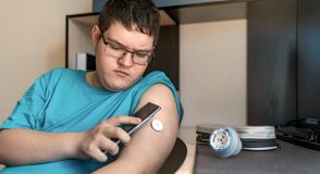 Ein Jugendlicher mit Brille betrachtet auf einem Smartphone seinen Glukosesensor am Oberarm. Auf dem Tisch liegen weitere Diabetes-Utensilien.