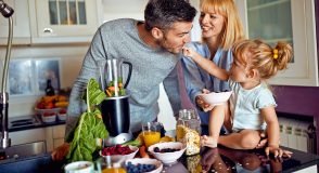 Eine Familie bereitet in der Küche gemeinsam ein ballaststoffreiches Frühstück zu. Ein Mann mixt grünes Blattgemüse und Karotten, während eine Frau und ein Mädchen mit Schalen voller Joghurt, Beeren und Haferflocken danebenstehen.