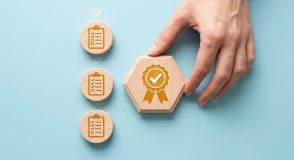 Eine Hand platziert einen sechseckigen Holzbaustein mit einem Häkchen und einem Auszeichnungs-Symbol neben drei runden Holzplättchen mit Checklisten.