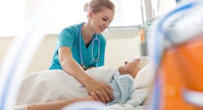 Eine junge Krankenschwester kümmert sich um eine ältere Frau in einem Krankenhausbett. Beide lächeln sich an.
