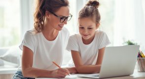 Eine Frau mit Brille und ein Mädchen sitzen gemeinsam an einem Tisch vor einem Laptop. Die Frau macht sich Notizen, während beide auf den Bildschirm schauen.