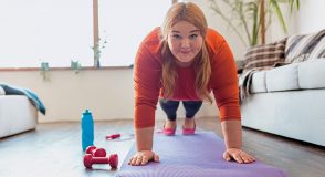 Eine Frau macht auf einer Trainingsmatte eine Plank-Übung in einem Wohnzimmer. Neben ihr liegen rote Hanteln und eine blaue Trinkflasche.