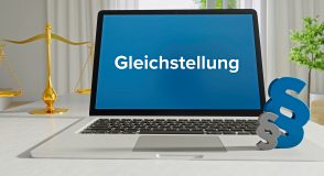 Laptop mit blauem Bildschirm und der Aufschrift „Gleichstellung“; daneben stehen eine goldene Waage und ein Paragraphenzeichen als Symbole für Recht und Gesetz.