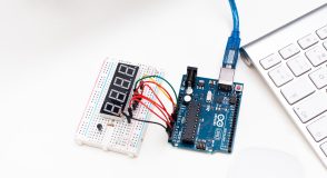 Ein Steckbrett mit Siebensegmentanzeige und farbigen Kabeln ist mit einer Arduino-Platine verbunden, die per USB-Kabel an eine Computertastatur angeschlossen ist. Sinnbild für selbstgebaute Open-Source-Elektronik, wie sie bei DIY-Systemen zur automatisierten Insulindosierung zum Einsatz kommt.
