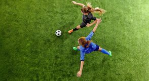 Zwei Fußballspielerinnen in schwarzen und blauen Trikots laufen auf einem grünen Rasen einem Fußball hinterher. Die Aufnahme aus der Vogelperspektive symbolisiert Bewegung und Prävention.