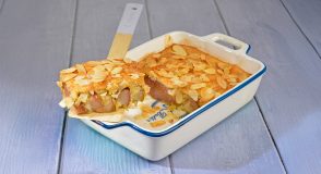 Mandel-Birnen-Auflauf in einer weiß-blauen Auflaufform, goldbraun gebacken und mit Mandelblättchen bestreut, ein angeschnittenes Stück mit sichtbaren Birnenstücken liegt auf einem Holzspatel.