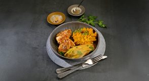 Gebratene Putenfilets mit Kürbispüree und gebratenem Chicorée, mit Petersilie bestreut, in einer Schale auf einem dunklen Tisch angerichtet. Daneben liegen Besteck und kleine Schälchen mit Gewürzen.