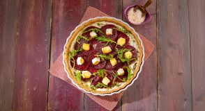 Rote-Bete-Tarte in einer weißen Tarte-Form, belegt mit dünnen Rote-Bete-Scheiben, Camembert-Stücken und Rucola, auf einem rosa Tuch und einem Holztisch, daneben eine kleine lila Schale mit grobem Salz.