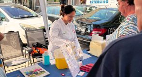 An einem Informationsstand unter einem Pavillon berät eine Frau im weißen Kittel eine Besucherin. Auf dem Tisch liegen Broschüren und Materialien zur Blutzuckermessung.