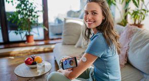 Junge Frau mit Glukosesensor am Oberarm hält ein Smartphone mit geöffneter Diabetes-App in den Händen und lächelt in die Kamera. Dargestellt wird die digitale Blutzuckerkontrolle im Alltag.