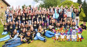 Großes Gruppenfoto auf einer Wiese: Zahlreiche Kinder, Jugendliche und Eltern in schwarzen T-Shirts mit der Aufschrift „Gemeinsam löwenstark!“ strecken jubelnd die Arme in die Höhe, rechts unten sind bunte Figuren von Fantasietieren eingeblendet.
