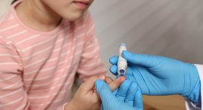 Eine medizinische Fachkraft mit blauen Einmalhandschuhen nutzt an einem Finger eines Mädchens in rosafarbenem Ringelshirt eine Stechhilfe, um Blut für die Glukosemessung zu gewinnen.