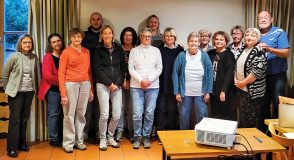 Gruppenfoto von 15 Personen in einem Vereinsraum nach einem Vortrag, im Vordergrund ein Beamer auf einem Tisch. Referentin Cornelia Sichermann steht als vierte Person von rechts in der Gruppe.